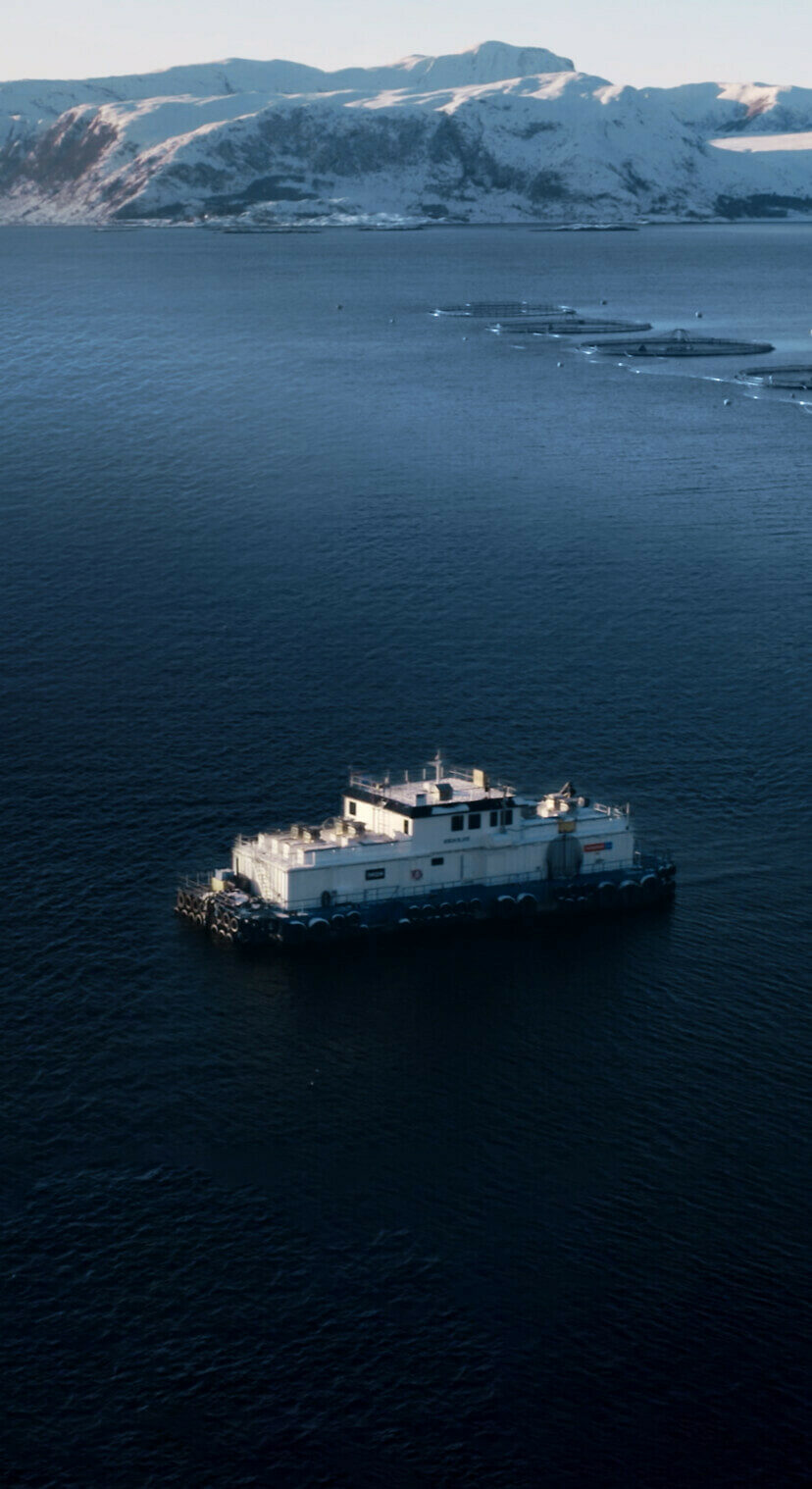 A white barge floats on dark blue water, with snow-capped mountains and fish farms in the distance.