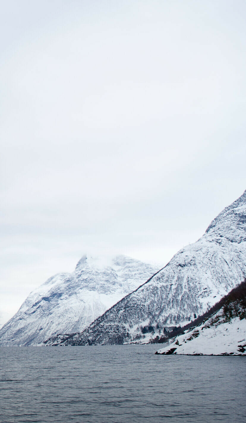 Snow-covered mountains rise above a dark, rippling body of water under an overcast sky.