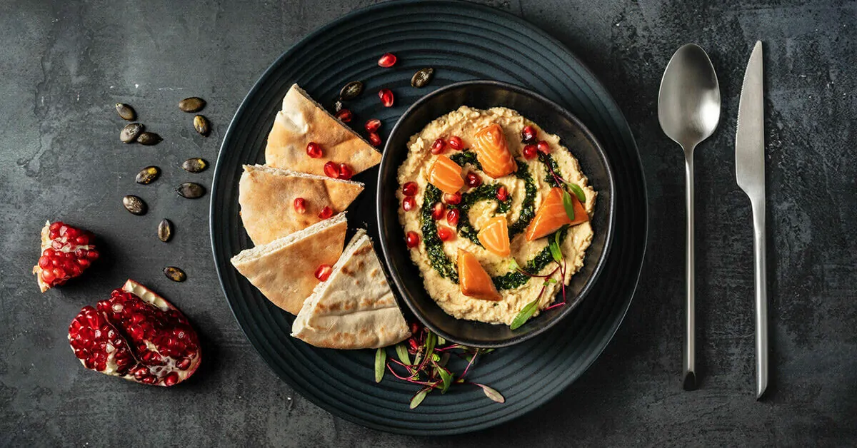 Hummus with salmon, pomegranate, and pita bread on a dark plate with cutlery.