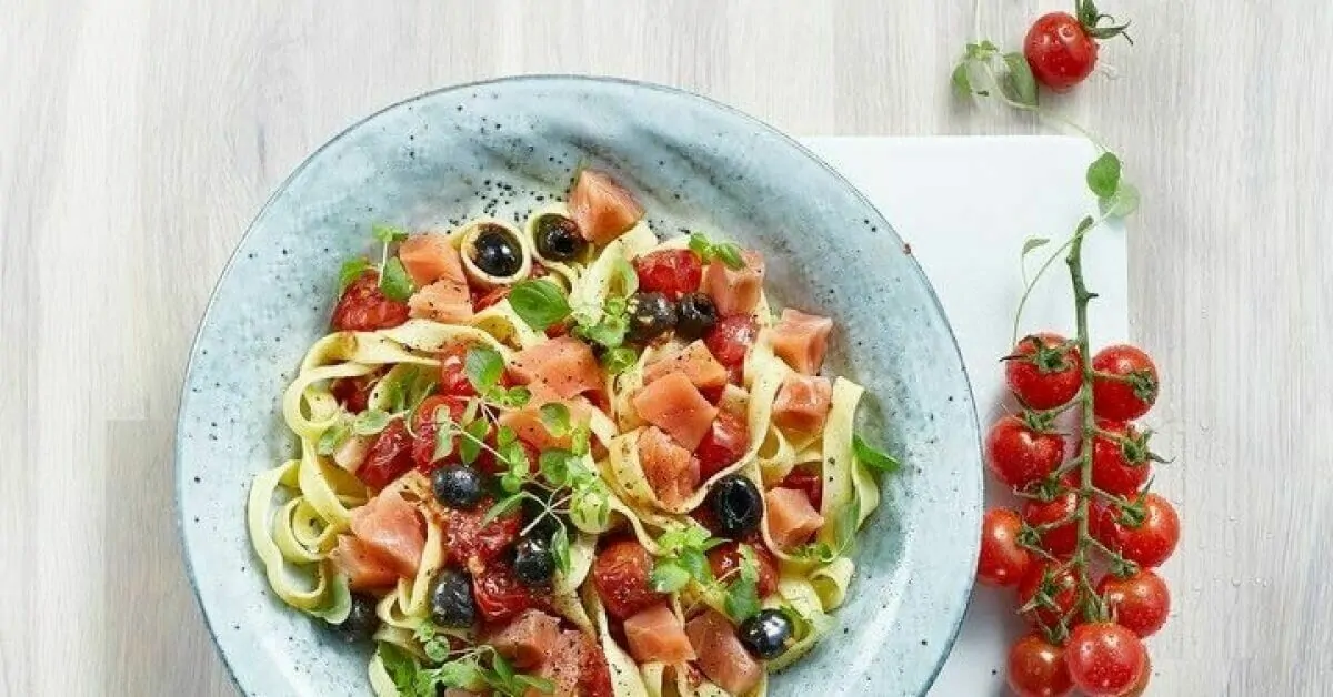 Fettuccine pasta with salmon, tomatoes, olives, and herbs on a blue plate.