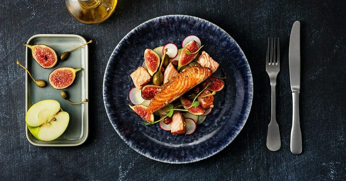 Salmon dish with figs, radishes, capers, and apple slices, served with cutlery.