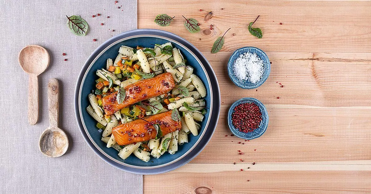 A plate of pasta with salmon, vegetables, and herbs, served with two small bowls of spices.