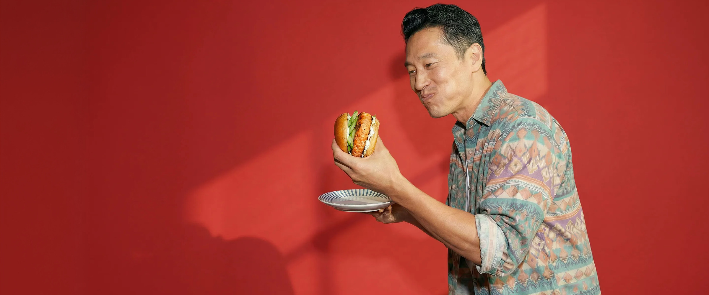 Man in a colorful shirt holds a burger against a red background.
